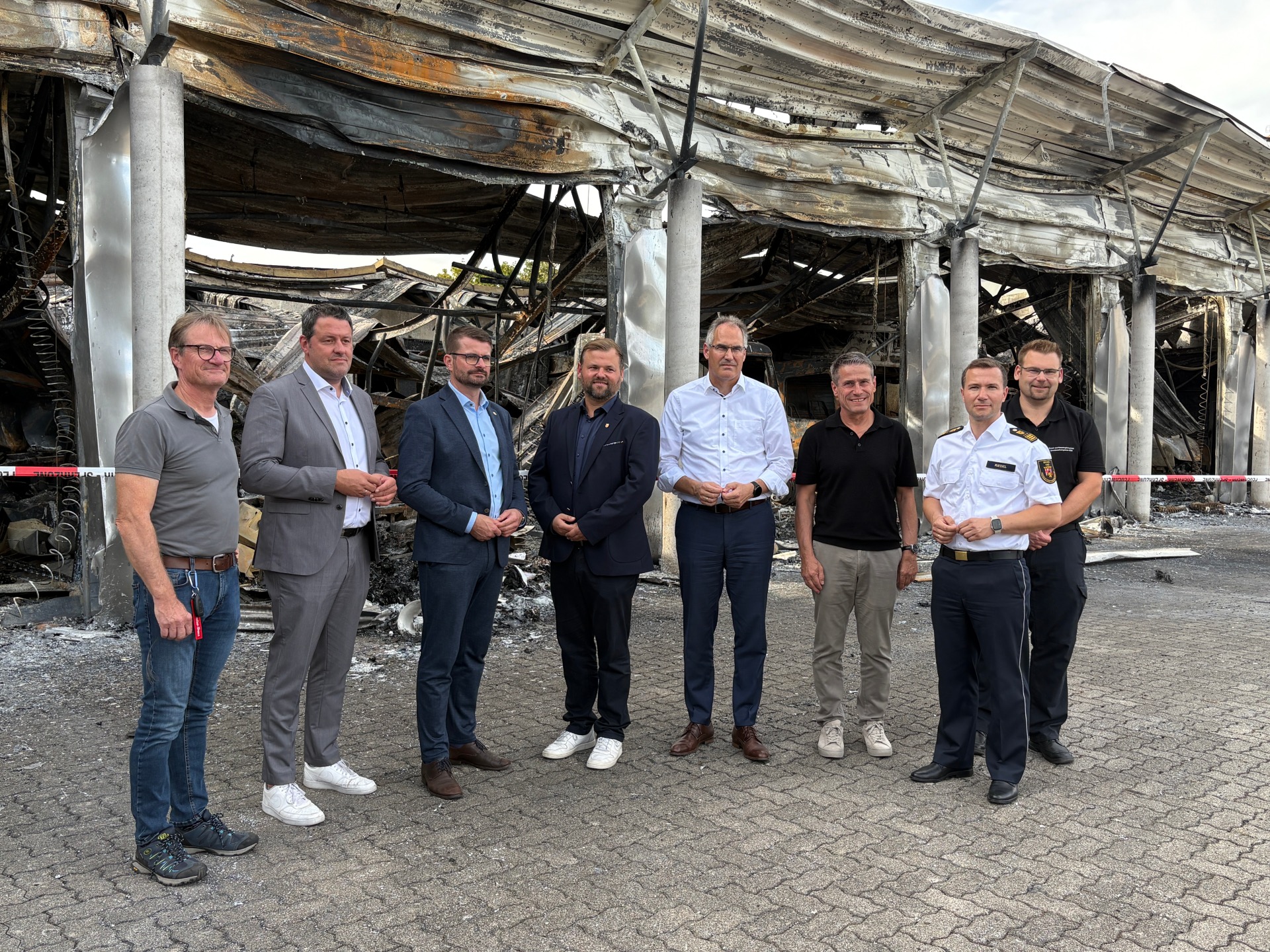 vlnr: Oliver Siebert (Wehrleiter VGO), MdL Tobias Baumgärtner, Staatssekretär Dirk Herber, Bürgermeister Simon Wingerter, Landrat Dietmar Seefeldt, Uwe Huth (Kreisbeigeordneter SÜW), Tom Riedel (LBKI), Jens Thiele (BKI SÜW) vor der ausgebrandten Feuerwehrhalle