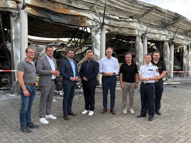 vlnr: Oliver Siebert (Wehrleiter VGO), MdL Tobias Baumgärtner, Staatssekretär Dirk Herber, Bürgermeister Simon Wingerter, Landrat Dietmar Seefeldt, Uwe Huth (Kreisbeigeordneter SÜW), Tom Riedel (LBKI), Jens Thiele (BKI SÜW) vor der ausgebrandten Feuerwehrhalle